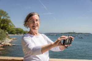 A woman of retirement age holds phone up to photograph something. Ocean water in the background.
