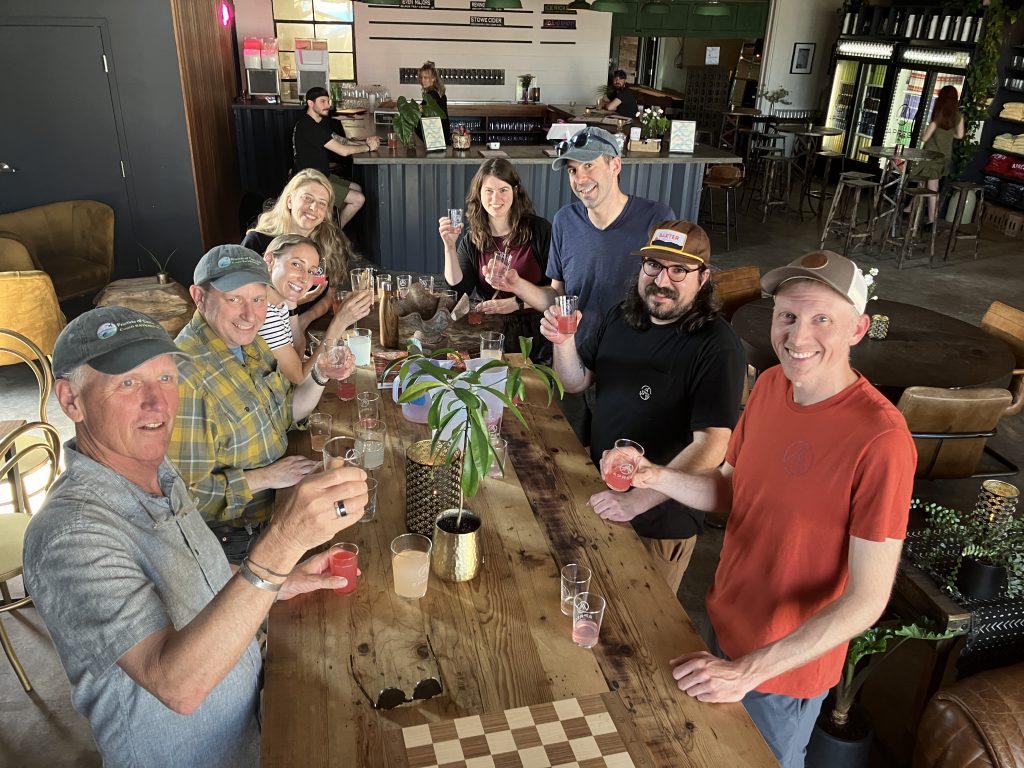 A group of people stand around a table, smiling, and holding up small clear glasses of pink colored drinks.