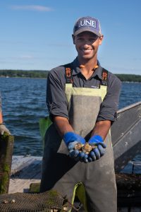 Michael holding oysters, wearing fishing gear, on a dock by the water