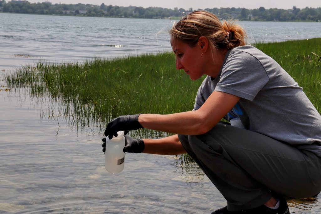Heather kneels beside the shore on Back Cove gathering water for PFAS testing into a container.