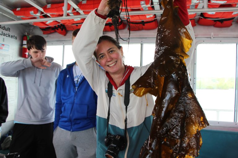 Meghan holds up a large piece of sugar kelp from inside the boat, a camera around her neck.