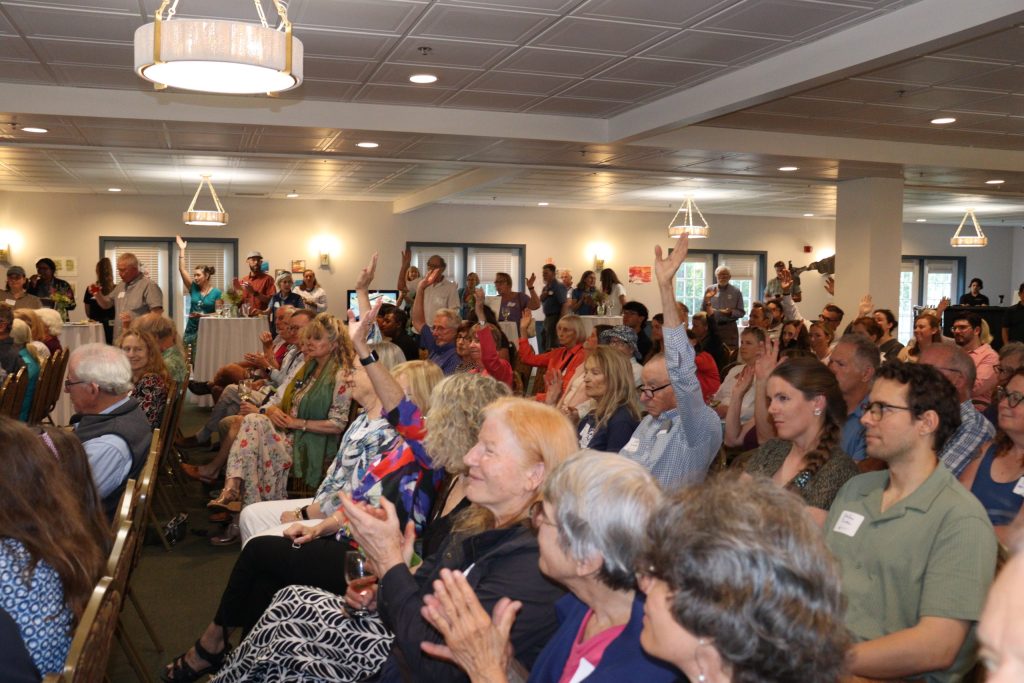 a large group sits for a meeting. Several people have their hands raised in the air.