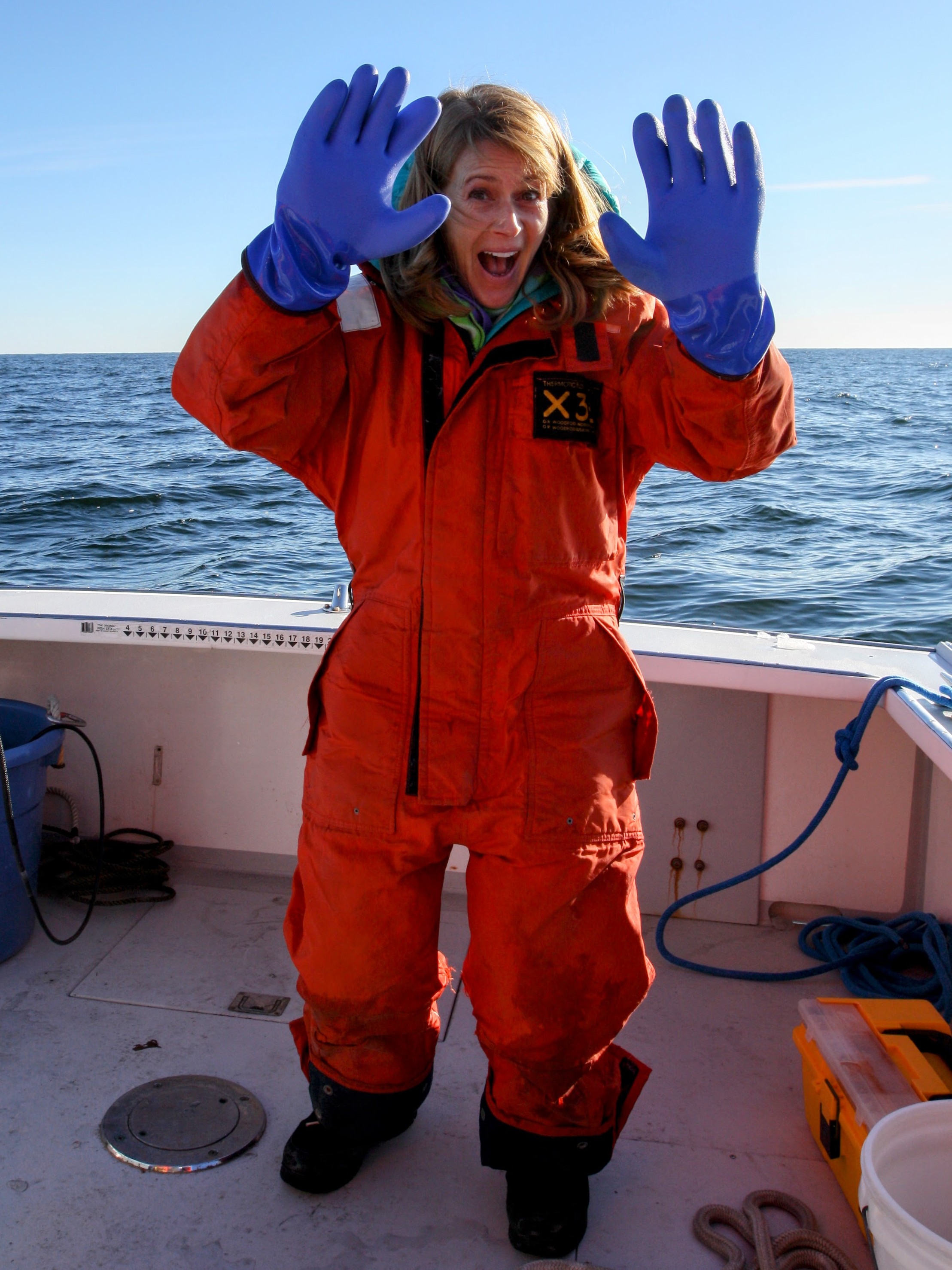 Ivy in an orange survival suit, holding up her hands in huge blue gloves